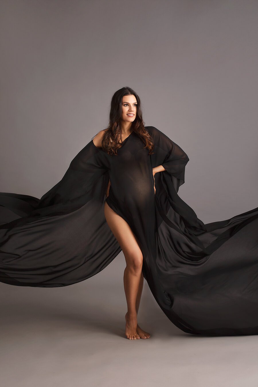 Brunette pregnant model wears a long chiffon cape in black color. She holds her waist with both hands. The cape draping fabric has been throw before the close up, so in the photo looks like it is flowing,