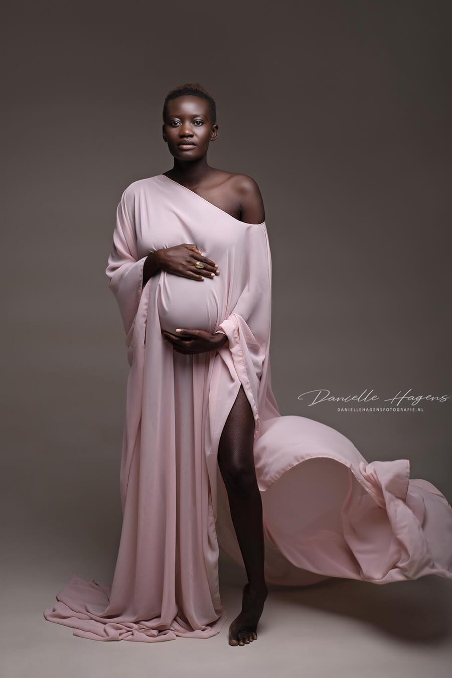 Pregnant woman poses in a studio with a chiffon cape in dusty pink color. She stares at the camera and holds her bump with both hands.