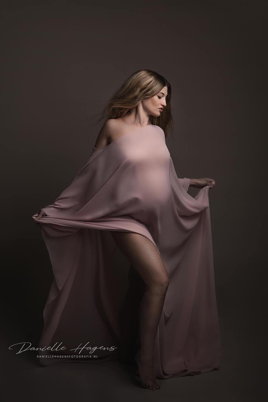 Blond pregnant model poses in a studio with a long chiffon cape made of chiffon. She holds the train in a playful way and faces down. 