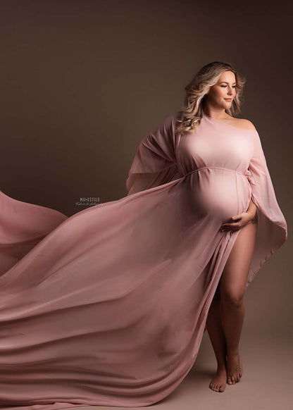 Blond pregnant model is wearing a cape made of chiffon in Dusty Pink color. One hand is under her belly and the other one is in her side. She is looking to the side with her eyes down. The ends of the cape are tossed to the side.