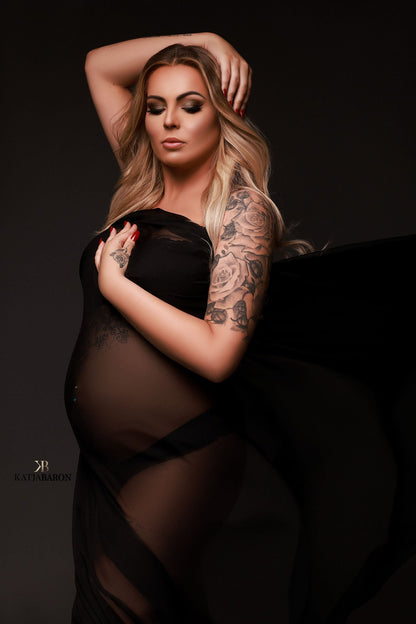 A blonde pregnant woman is covered in black chiffon fabric. She has long blonde hair and has a flower tattoo on her upper arm. The photo has a chique effect.