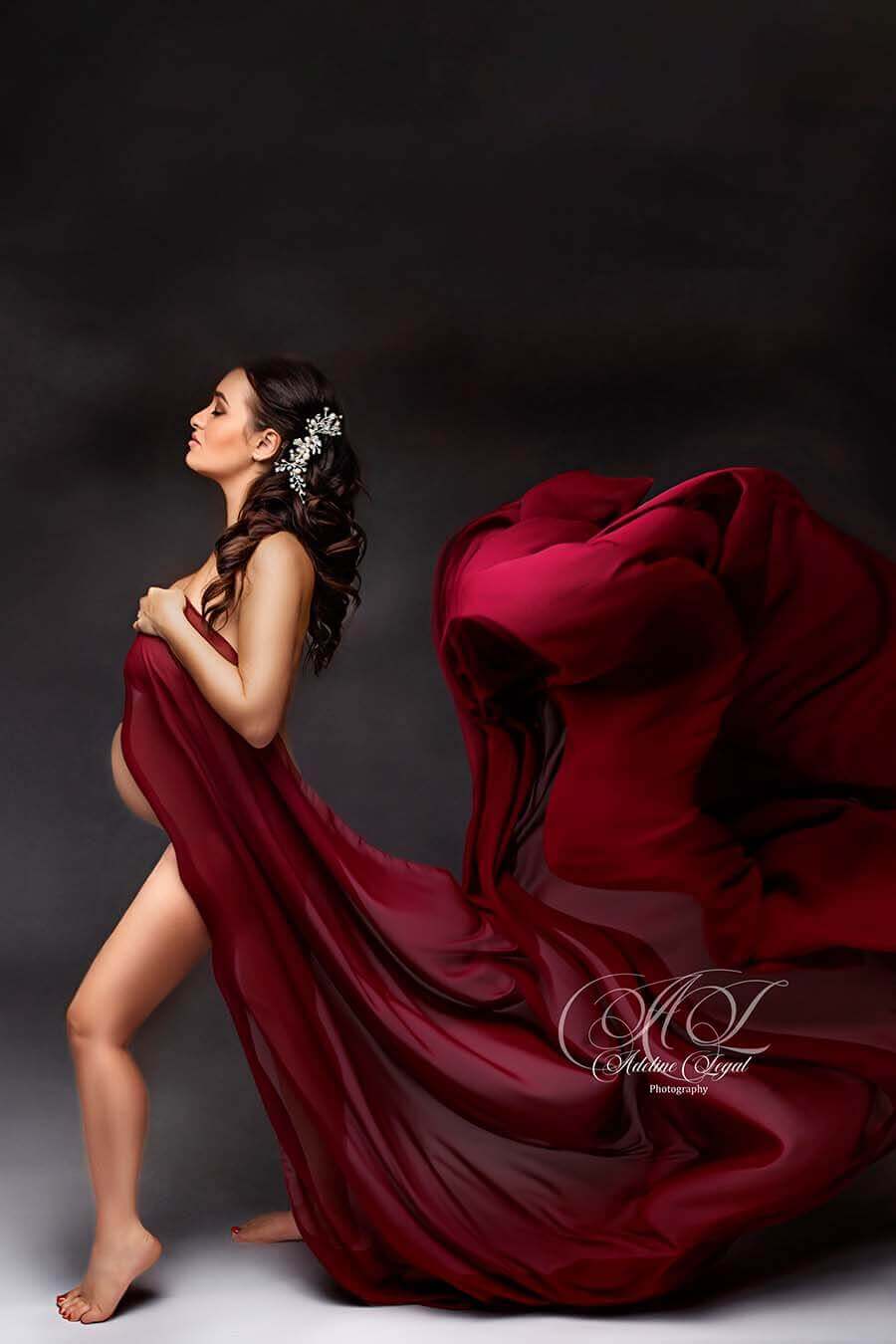 brunette pregnant model poses in a studio on her profile, face facing up with her eyes closed. she is wearing a chiffon draping fabric in bordeaux color. she has a pretty sparkling accessory on her head. she is holding the chiffon fabric against her chest and by this creates a flowing movement with the light fabric. 