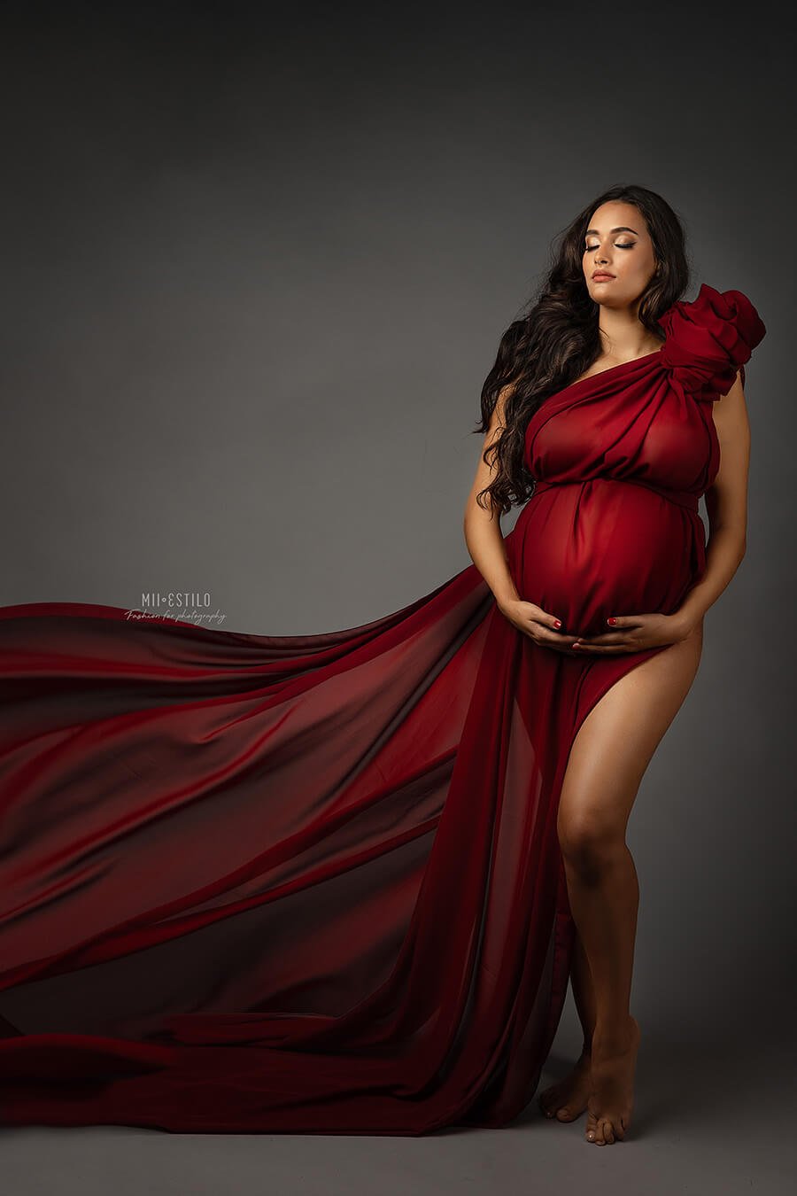 brunette pregnant model poses with her eyes closed in a studio wearing a chiffon bordeaux scarf tied as a one shoulder dress.