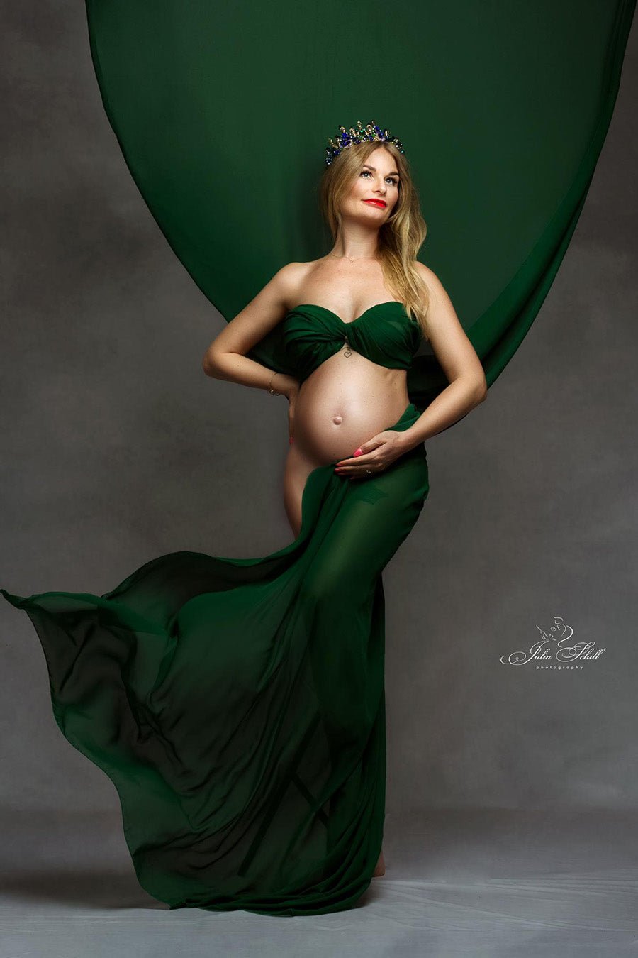 tall blond pregnant model poses in a studio wearing a chiffon scarf in dark green color wrapped around her body. she has a crown to match the look.