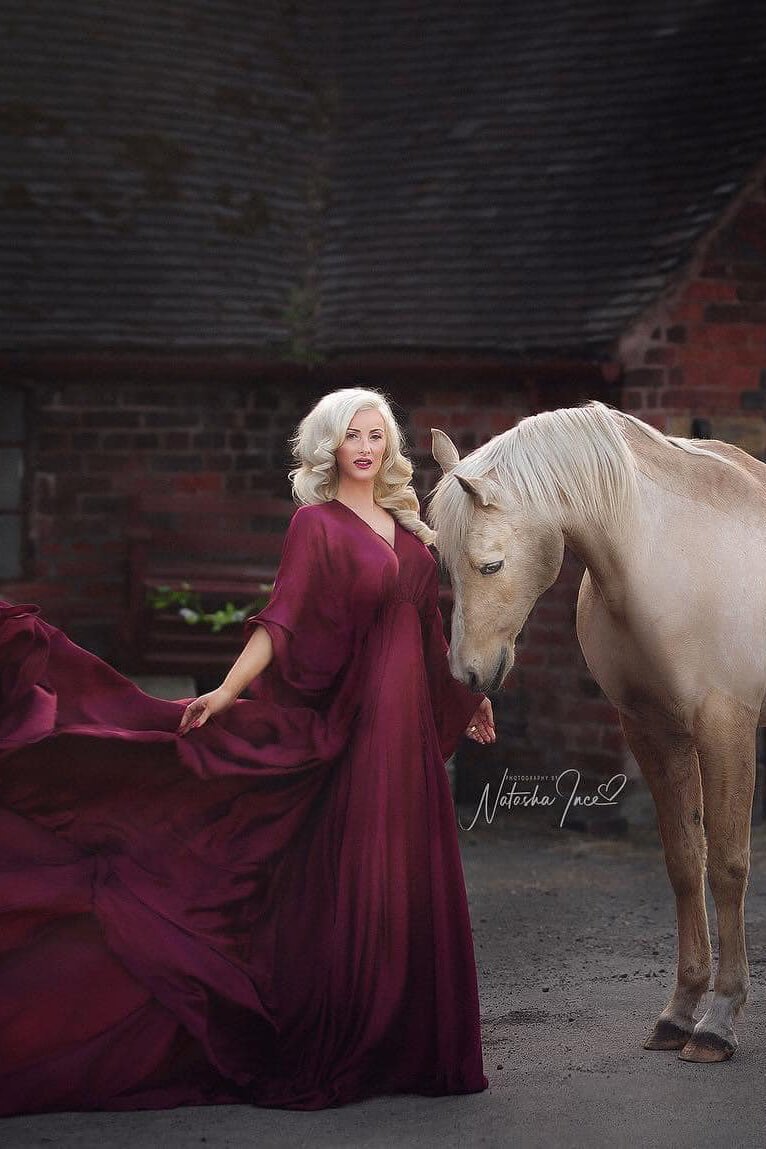 A woman is posing in a long dress with her horse next to her. The dress is being tossed behind the woman. The dress has a v neckline and 3/4 sleeves. 