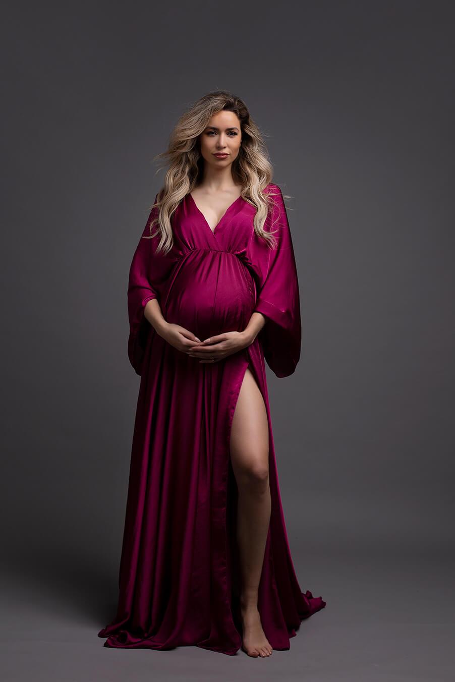 The woman in this photograph is pregnant. She has long blonde hair. The dress that she is wearing is long and in the color cherry. The dress has a v-neckline 