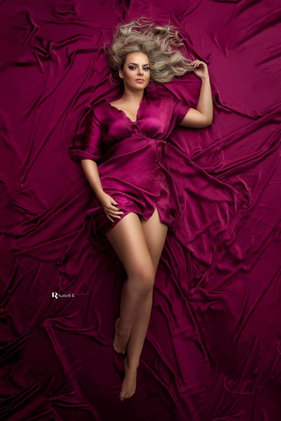 A model is laying on the ground. She is wearing a dress with a lot of fabric. The floor around her is covered in the same fabric. The color of the dress is cherry and the material is shiny. 