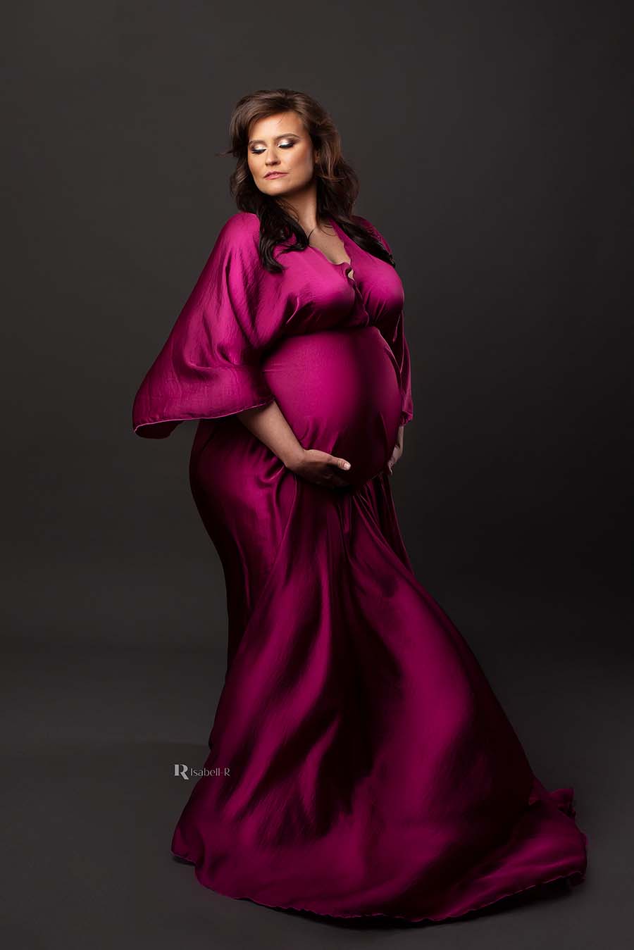 The pregnant model is in a studio. She is wearing a wide boho fit dress. The dress is in the color cherry and has a silky material for a little shine. The dress is moving in the wind.