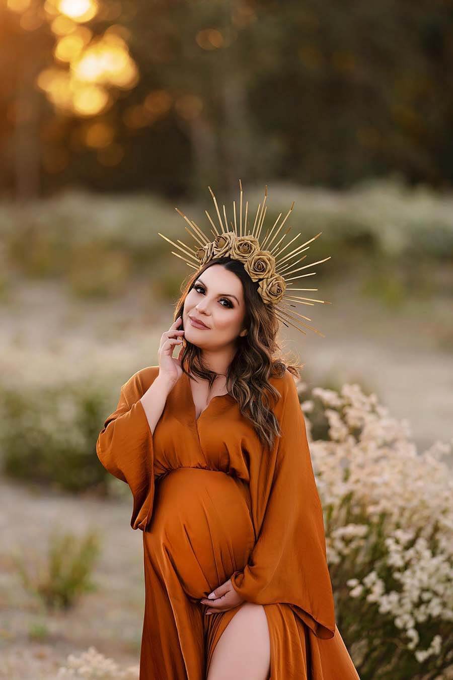 Pregnant woman poses outside in front of some wild white flowers. She is wearing a cognac silky dress with a low v cut neckline. One hand holds her bump and the other touches her face. One of her legs can be seen through the split on the dress and she has a crown with flowers to match the style. 