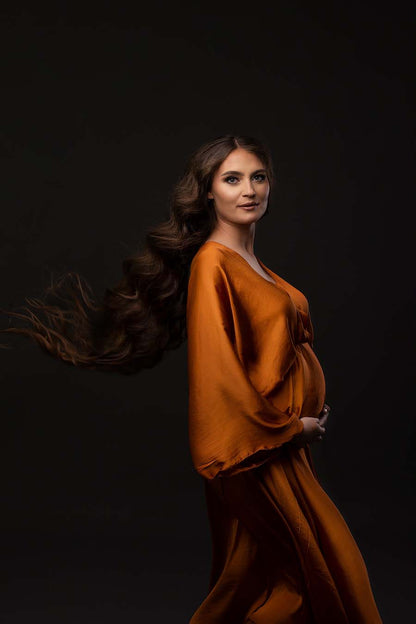 Pregnant model poses in a studio wearing a long dress made of silk in cognac color. She is posing on her side and holds her bump with both hands. She stares at the camera. The dress has long kaftan style sleeves. 