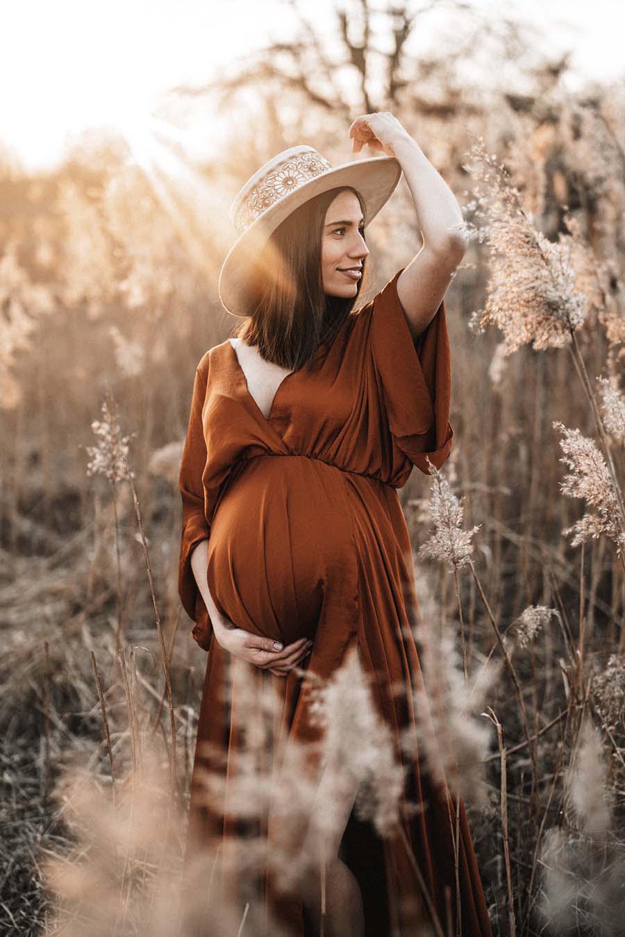 brunette model with short hair poses outside in a flower field for her maternity photoshoot. she has a matching hat that matches perfectly the bohemian style form the photo. she is wearing a long cognac dress with kaftan style sleeves and a low v cut neckline.