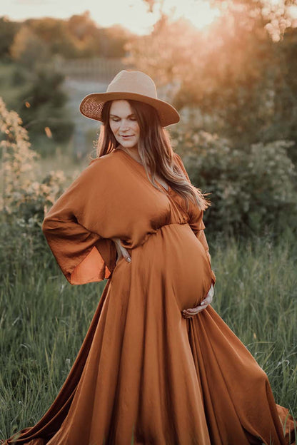 Pregnant woman poses outside wearing a cognac long maternity dress made of silk. The dress features kaftan style sleeves and a long circle skirt. She wears a hat to match the style.