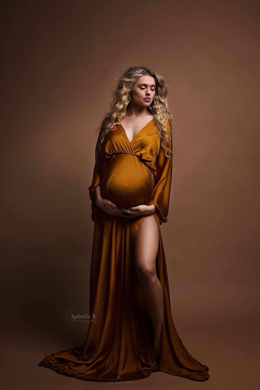 Model is wearing a long dress with a split by the leg. The dress is boho styled and in the colour cognac. The woman is pregnant and holding her belly