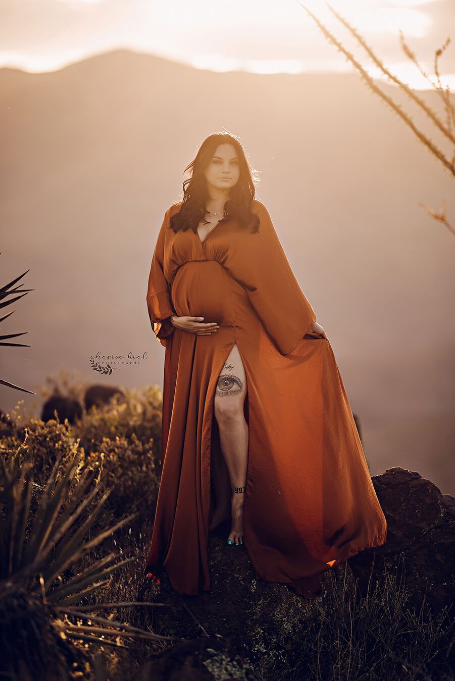 The woman is standing outside with sunlight behind her. She is wearing a long dress in the color cognac. The dress is boho styled and she is holding it with her hand. The other hand is by her belly because she is pregnant