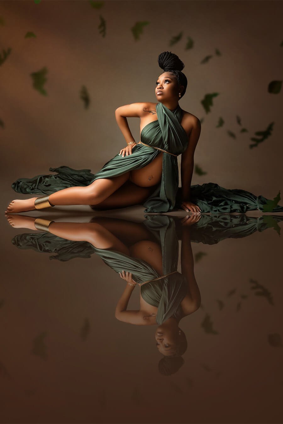 Pregnant woman with black braids in a knot poses in a photography studio. She sits on the ground with on arm holding her up and the other one in her side. A silky scarf is wrapped around her with a sash around her waist.