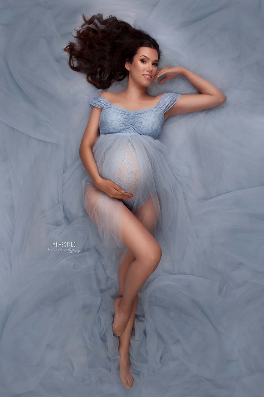 brunette model lays on the floor of a studio during her maternity photo shoot. she wears a bodysuit made of lace with a sweetheart neckline and drop sleeves. the bodysuit has a long tulle skirt attached and the model is laying in the tulle. the whole set is light blue.