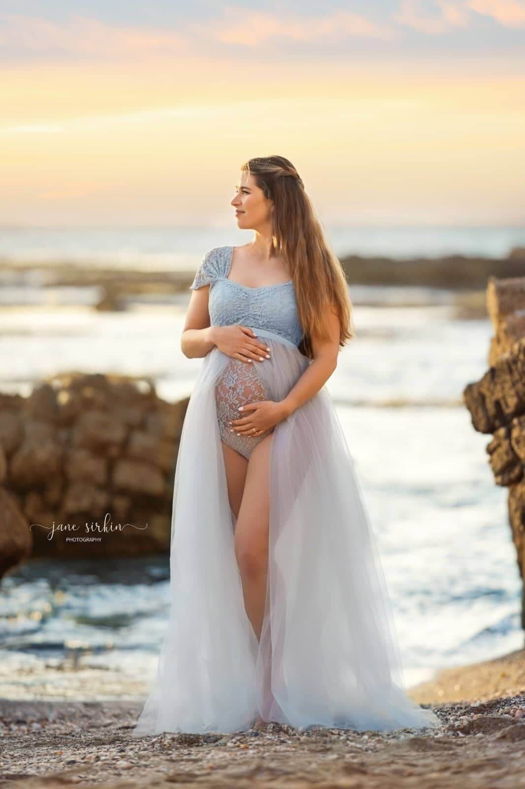 blond pregnant model poses outside by the water wearing a maternity dress in light blue color. she is looking to the side and holds her bump with both hands. the dress is a bodysuit made of lace with a tulle skirt attached. the skirt has an opening in the middle where her legs can be seen. 