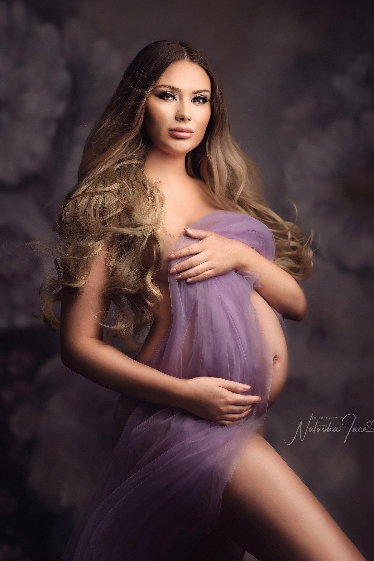 model poses in a studio for a maternity photoshoot wearing a tulle draping fabric in purple color.