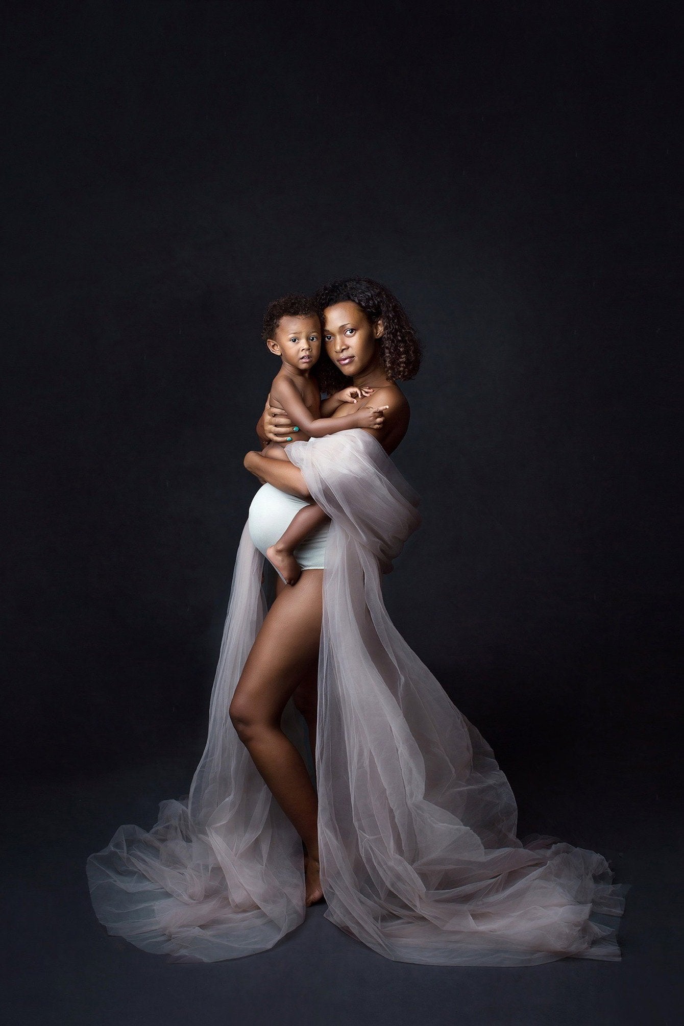 model poses in a studio for a maternity photoshoot wearing a tulle draping fabric in white color. she is holding her baby and wearing an off white strapless bodysuit underneath.