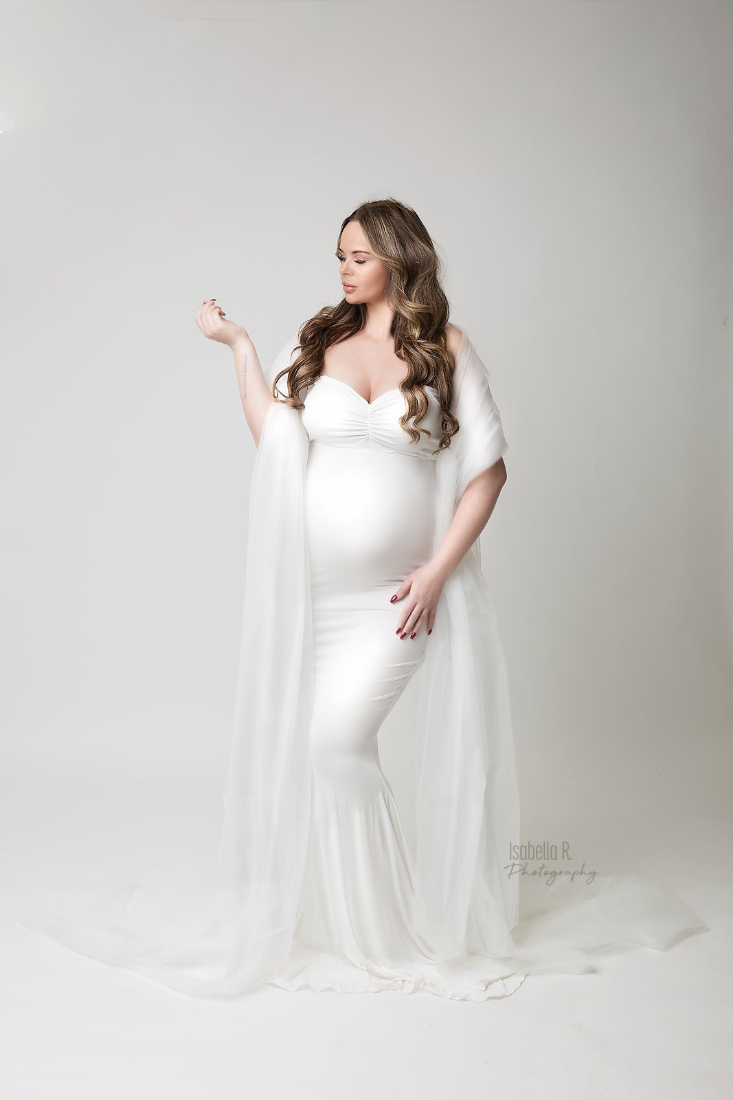 model poses in a studio for a maternity photoshoot wearing a tulle draping fabric in white color. she is wearing a strapless tight white dress.