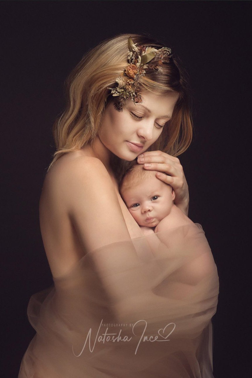 model poses in a studio for a maternity photoshoot wearing a tulle draping fabric in sand color. she is holding her newborn close to her and has her eyes closed.