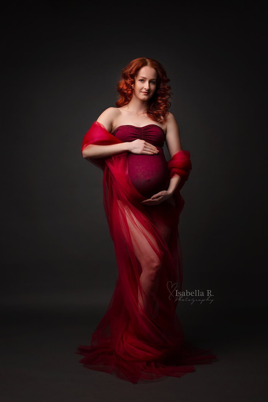 model poses in a studio for a maternity photoshoot wearing a tulle draping fabric in red color around her arms. she also wears a strapless red bodysuit.