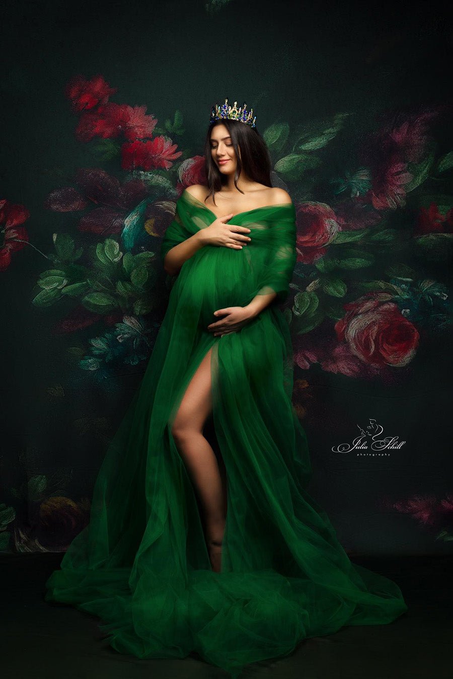 pregnant model poses in a studio against a flower background wearing a dark green tulle scarf covering her boddy. she has her eyes closed and a flower crown to match the style. 