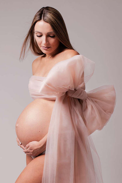 pregnant model poses with a tulle scarf tied as a bow covering her chest. She has her eyes partially closed and holds her bump gently with both hands.