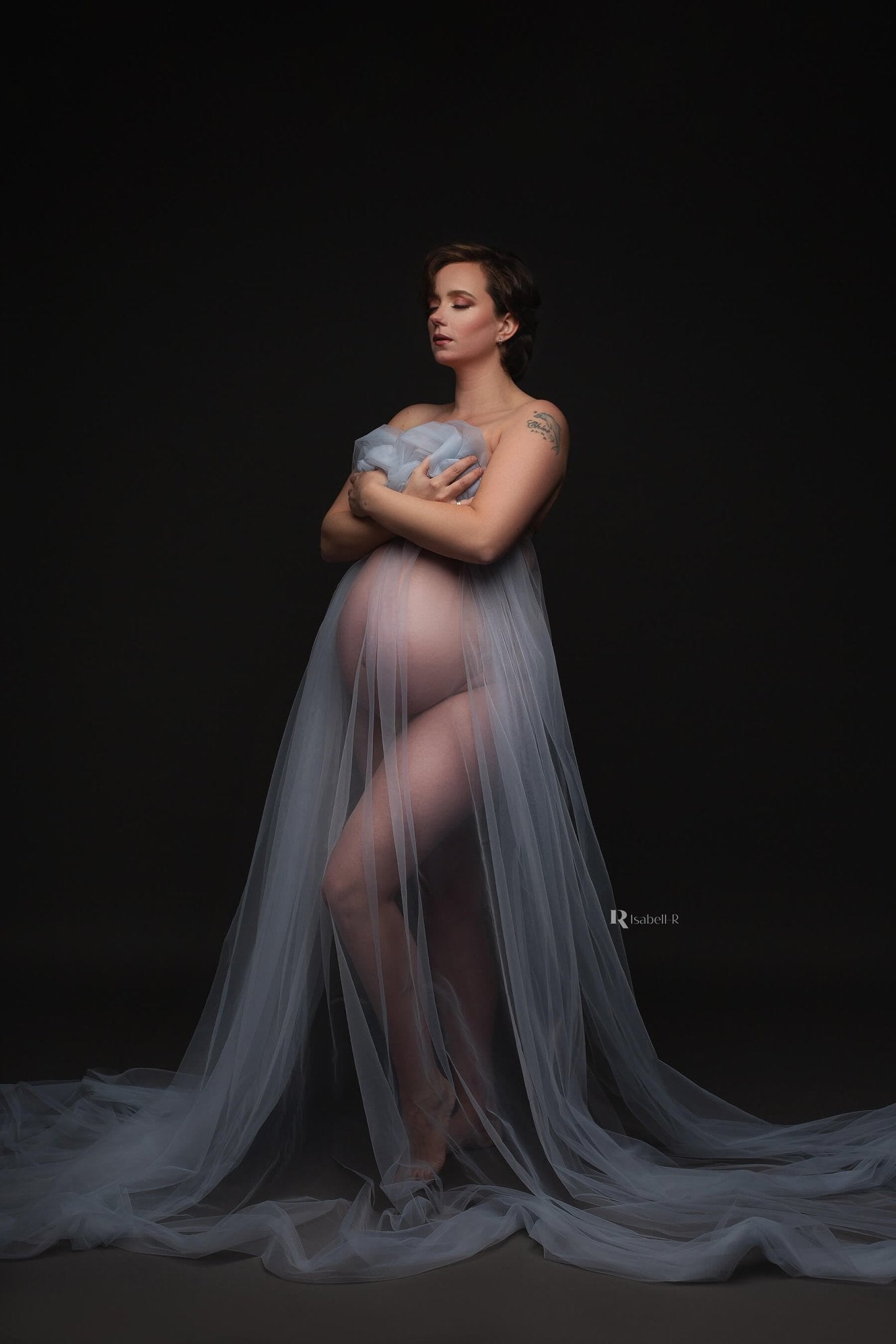 A pregnant model is using the tulle scarf light blue to cover up in the photo. she is holding the fabric up in front of her body with her hands. The rest of the fabric is going down to the floor . The look in this photo is dreamy.