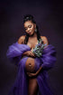 pregnant woman poses with a purple tulle scarf wrapped around her arms and holding a bouquet of lavenders. she is looking down and holds her bump with both hands. the scarf is purple. 