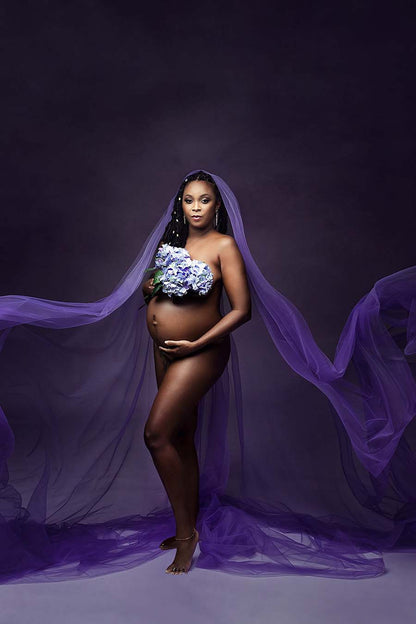 pregnant woman in a studio poses staring to the camera. she has no clothes, but her chest is covered with a bouquet of purple flowers. she has a tulle scarf on her head, like a mantel. 