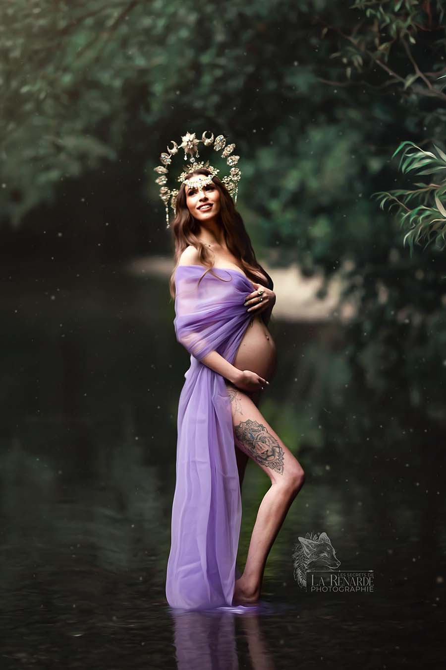 A pregnant woman is posing in a lake outside. She is standing in a little bit of water and is wearing the tulle scarf purple. She is holding the scarf as a robe with one hand. She is wearing a big headpiece to finish the look.