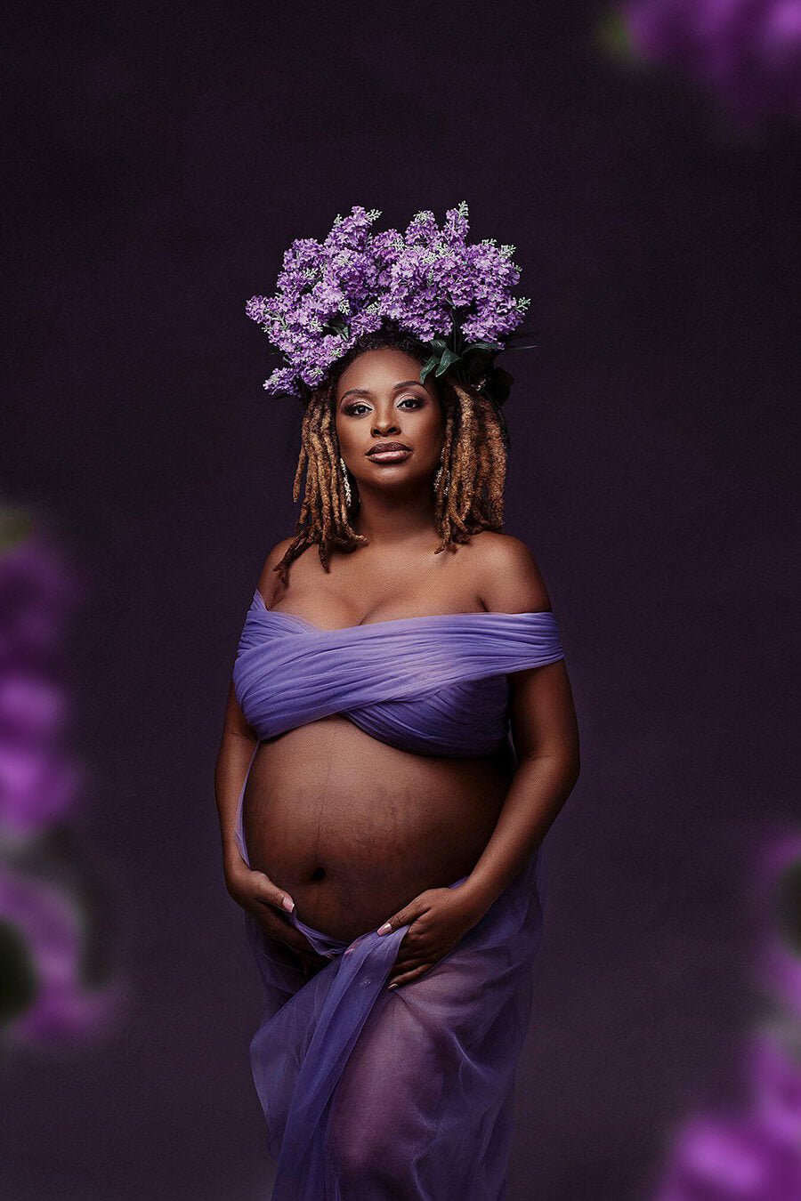 pregnant model poses in a studio wearing a tulle scarf tied around her breasts and the rest covering her upper legs and waist, like a skirt. She is staring to the camera and has a flower crown on her head in purple color, to match with the scarf that is also purple.