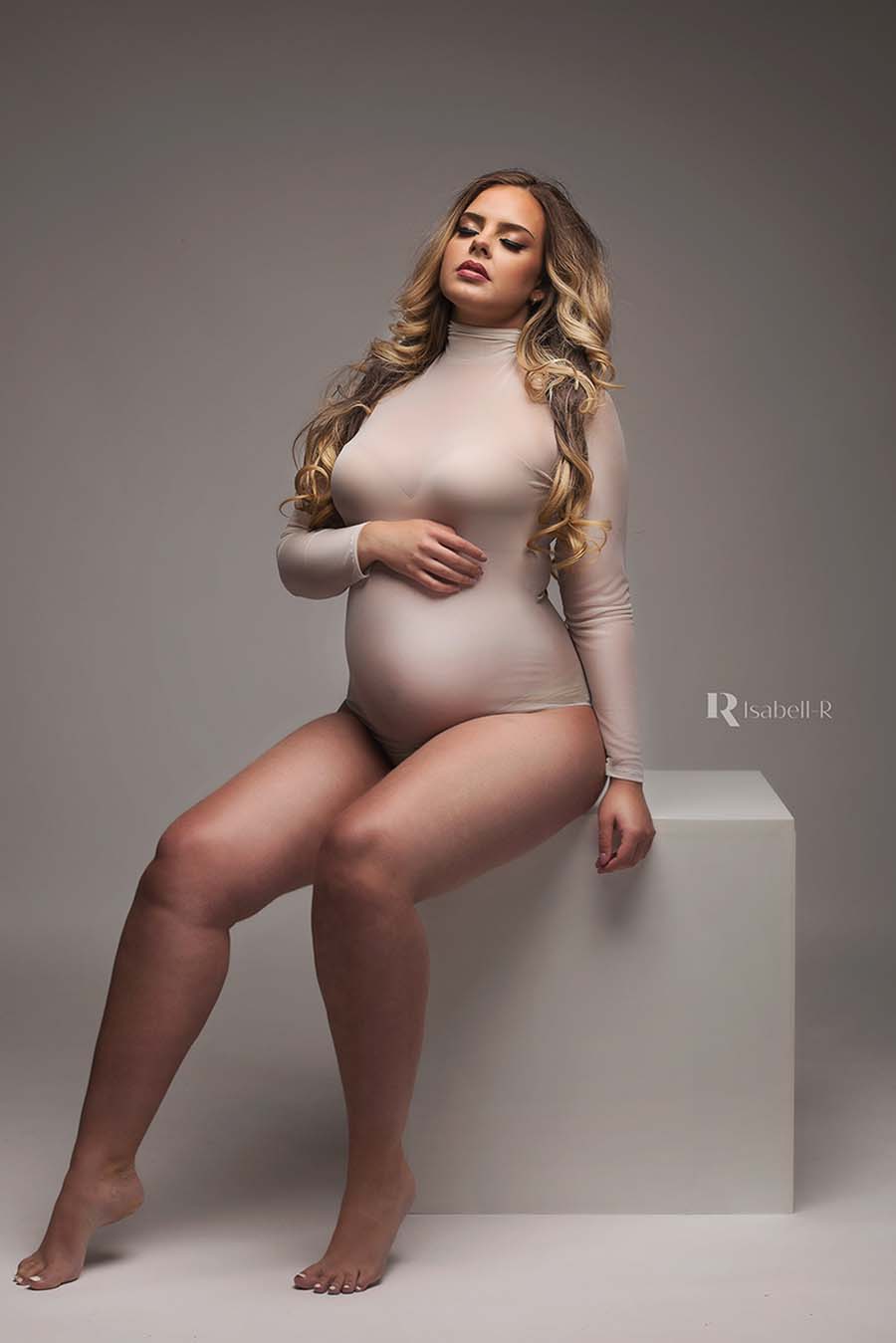 Blond long haired pregnant model poses in a studio during a maternity photography session. She is wearing an off white maternity bodysuit. She is wearing her own bra since the bodysuit can be a bit see through. She has her eyes closed and faces up - one of her hands holds her bump.