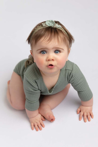 baby poses in a studio with a white background wearing an azur romper with long sleeves and a hair band.